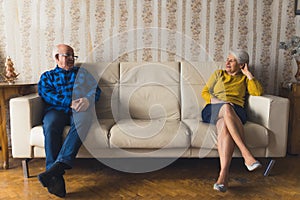 Elderly upset married couple sitting apart on leather sofa in their old-fashioned apartment after a quarrel.