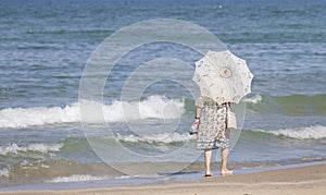 An elderly vintage lady with an openwork umbrella strolls along the beach