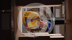 Elderly warehouse worker preparing goods for shipment while standing with clipboard near shelves with parcels ready for