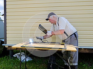 An elderly welder works with electric arc welding in the yard of a country house