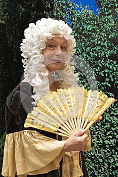 Elderly woman 60-70 years old in wig and old dress holding fan in her hand on background of greenery, 18th century clothes,