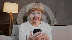 An elderly woman of 60s is sitting on the sofa with a phone in her hands