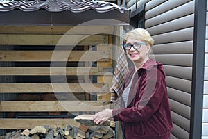 An elderly woman in a bard jacket is holding firewood for the stove.