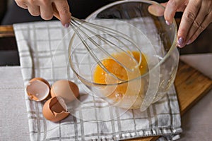 Elderly woman beats eggs with a wisk in a glass bowl. Prepare for making christmas pie or pancakes on kitchen table.
