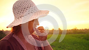 An elderly woman in a brown T-shirt and a white hat is trying to taste freshly raspberries on a sunset background, a