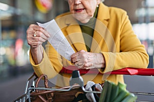 Elderly woman checking her receipt after purchase, looking at amount of money spent, ensuring all charges are correct.