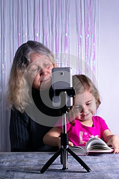 An elderly woman and a child are filming a video blog. Grandmother and granddaughter with a phone record video messages at home.