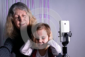 An elderly woman and a child are filming a video blog. Grandmother and granddaughter with a phone record video messages at home.