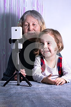 An elderly woman and a child are filming a video blog. Grandmother and granddaughter with a phone record video messages at home.