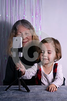 An elderly woman and a child are filming a video blog. Grandmother and granddaughter with a phone record video messages at home.