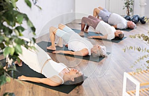 Elderly woman in company of hatha yoga lovers performs bridge pose, setu bandha sarvangasana