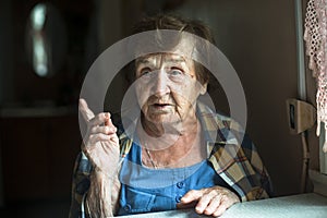 An elderly woman in a dark background near a window points her finger admonishingly.