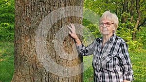 An elderly woman is depicted in the portrait, leaning against the trunk of a large tree, enjoying her walk in the woods.