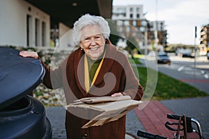 Elderly woman disposing cardboard into the garbage can, waste container in front of her apartment complex.