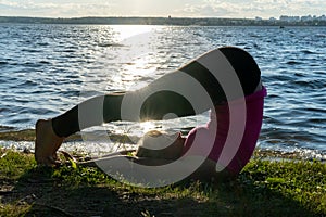 An elderly woman does sports at dawn on the shore of the lake. The woman throws her legs over her head