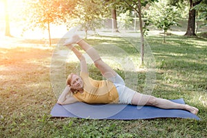 an elderly woman does yoga in the park and does complex stretching exercises by lifting her legs high up .