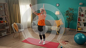 Elderly woman doing star jumps on a gym mat in the living room. A grayhaired retired woman takes care of her health