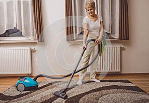 Elderly woman doing woman chores at home. Vacumming the carpet