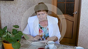 Elderly woman drinking tea at the table, watching pictures on the tablet.