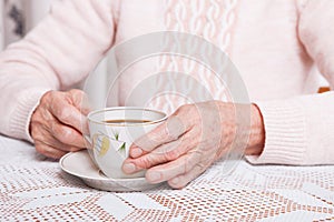 An elderly woman drinks tea at home. Senior woman holding cup of tea in their hands at table closeup