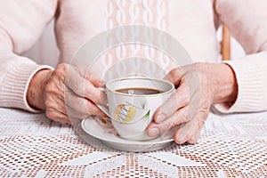 An elderly woman drinks tea at home. Senior woman holding cup of tea in their hands at table closeup