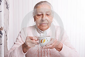 An elderly woman drinks tea at home. Senior woman holding cup of tea in their hands at table closeup