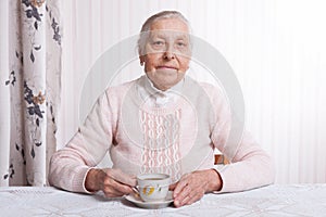 An elderly woman drinks tea at home. Senior woman holding cup of tea in their hands at table closeup