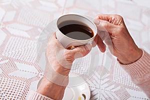 An elderly woman drinks tea at home. Senior woman holding cup of tea in their hands at table closeup. Horizontally top