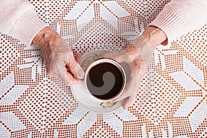 An elderly woman drinks tea at home. Senior woman holding cup of tea in their hands at table closeup. Horizontally top