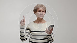 An elderly woman with a focused gaze holds and uses a smartphone, touches the screen and shakes wave index finger