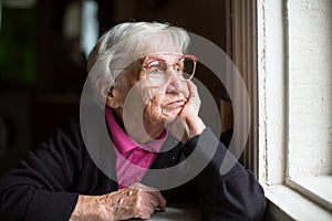 Elderly woman in glasses thoughtfully looking out the window. Loneliness.