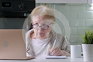 Elderly woman in glasses working using laptop at home, looking on computer