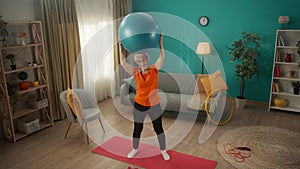 Elderly woman goes in for sports with a fitness ball in the living room. A greyhaired woman holds a ball over her head