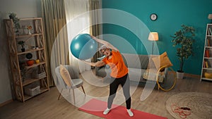 Elderly woman goes in for sports with a fitness ball in the living room. A greyhaired woman holds a ball over her head