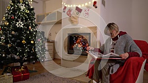 Elderly woman grandmother relaxing on a armchair in front of the family Christmas tree reading book. Fireplace