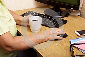 An elderly woman hand on a computer mouse. The old grandmother works behind the computer keyboard and drinks coffee