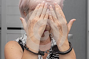 An elderly woman in handcuffs hides her face on a blurry background. Elderly criminal, criminal liability of pensioners, domestic