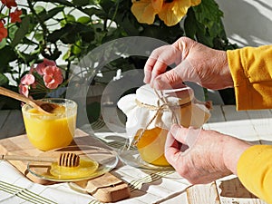 An elderly woman is hands wrap a glass jar of rustic natural flower honey. Honey and a cup of green tea on a background of flowers