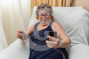 Elderly woman with headphones looking at smartphone in living room. Concept of older people using technology.