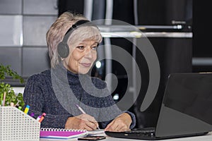 An elderly woman in her 60s and 65s is sitting in front of her laptop with headphones on. Concept: remote work, learning foreign l