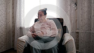 An elderly woman with her hobby sitting in a chair and knitting on the background of the window, looking with contempt