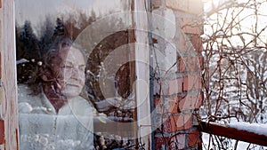 An elderly woman in her house looks out of a window with glare, her brick house on a winter garden in the light of the