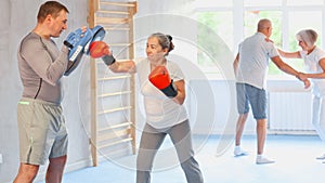 Elderly woman with her trainer at gym. Woman wearing boxing gloves exercise and punch to pads for boxing
