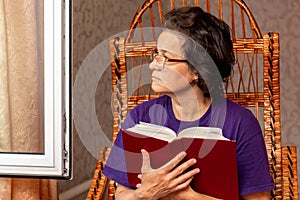 Elderly woman holding a Bible in her hand and looking out the open window, meditating on what she has read