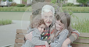 Elderly woman hugs and squeezes two young children as they sit and embrace on a park bench, they laughing and smiling