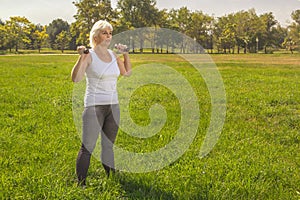 Elderly woman lifts dumbbells while doing fitness in a city park against the backdrop of mountains