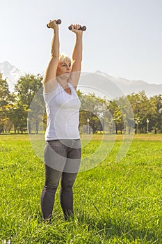 Elderly woman lifts dumbbells while doing fitness in a city park against the backdrop of mountains