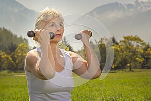 Elderly woman lifts dumbbells while doing fitness in a city park against the backdrop of mountains on a sunny day.