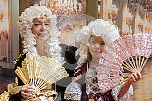 An elderly woman and a little girl dressed in elegant 18th century style clothing pose with hand fans at a costumed