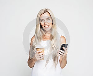 Elderly woman with long hair holding smartphone and takeaway coffee over grey color background